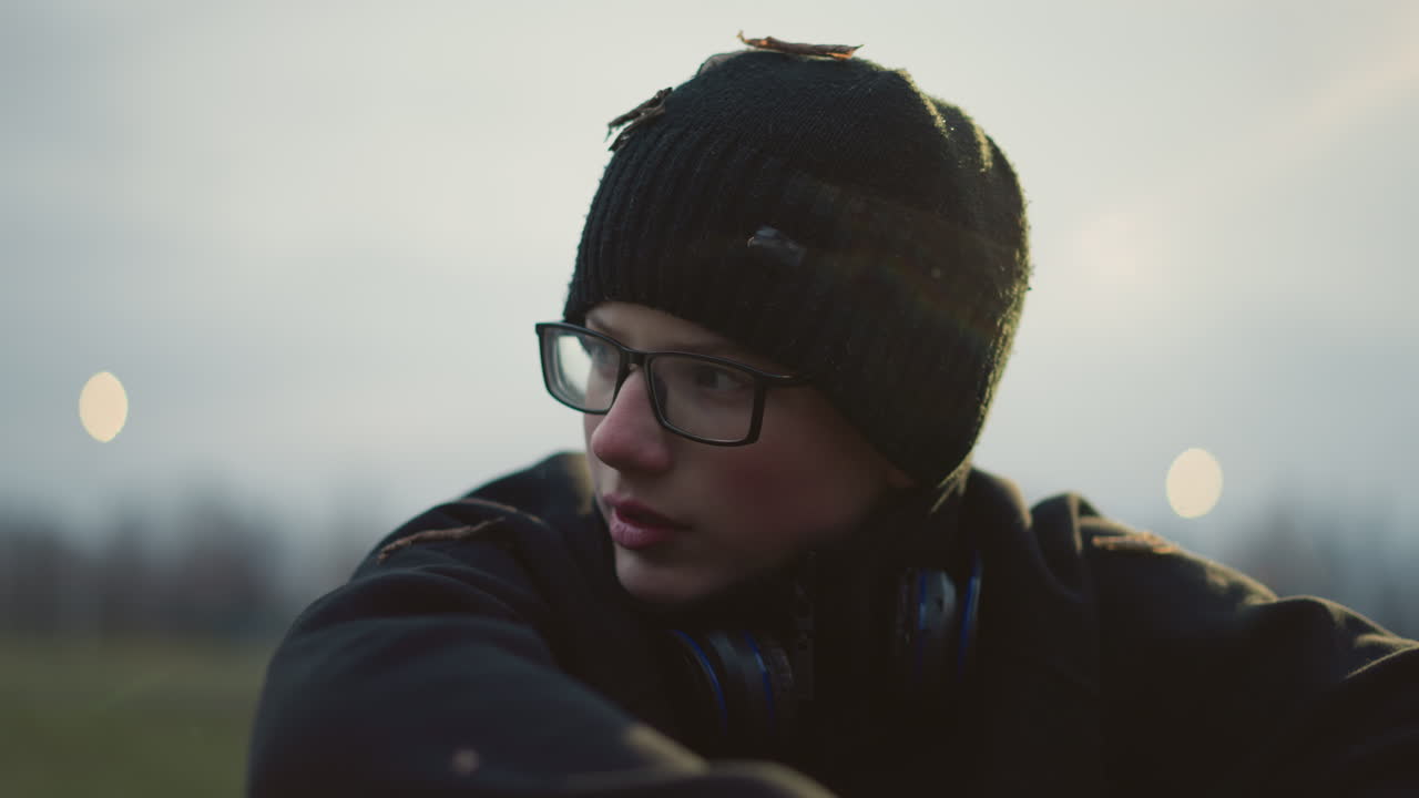 Close-up of a young boy wearing glasses and a black beanie with grass particles on his head and headphones around his neck, with light reflection on him