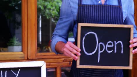 Smiling-waiter-standing-with-menu-and-open-sign-board