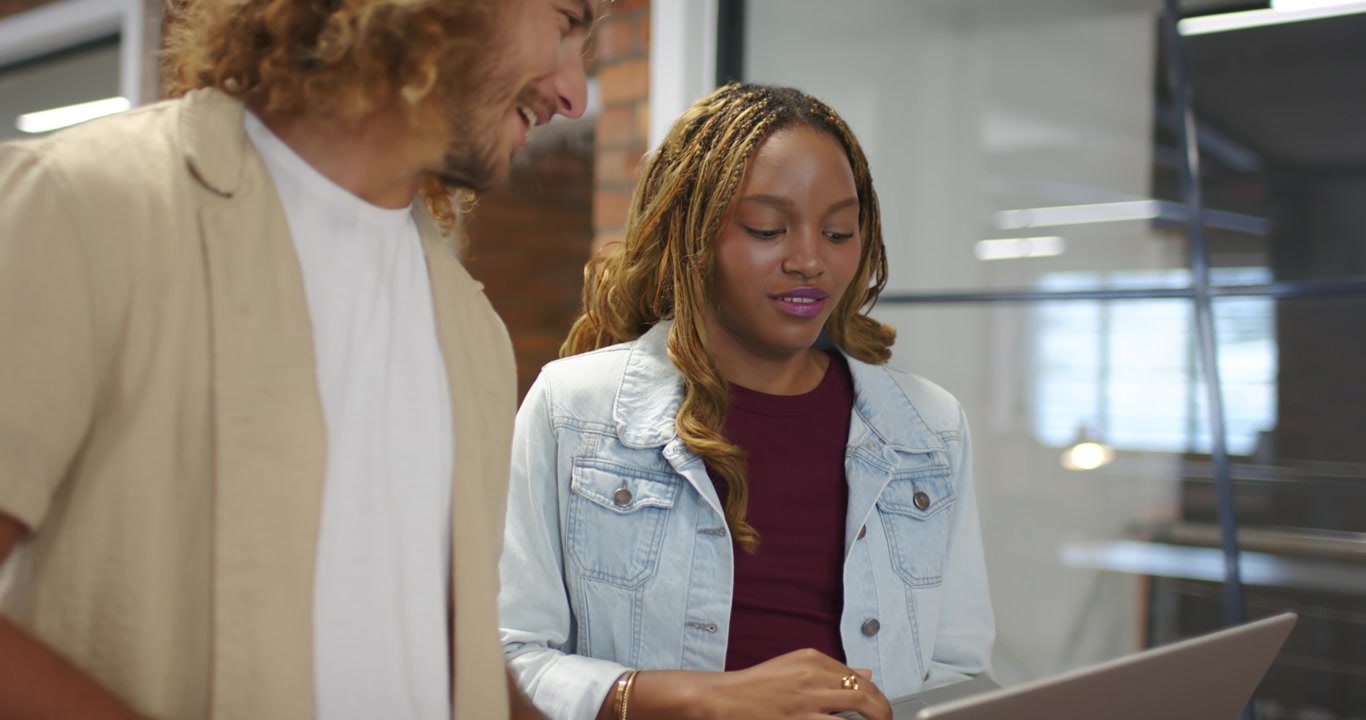 Colleagues at desk, woman in denim jacket navigating laptop reviewing content while man watching