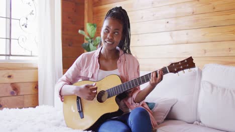 happy african american woman sitting on sofa and playing guitar in log cabin, slow motion