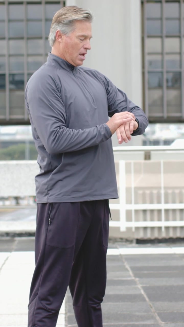 Vertical video: Glancing fit man in gray top checking watch on rooftop by railing, preparing to run