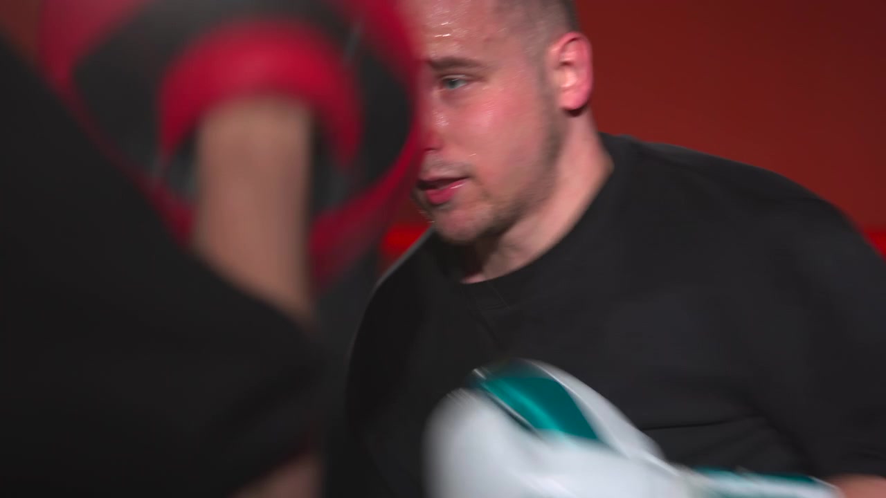 Giovane pugile all'interno di una palestra illuminata di rosso di fronte a un guantone bianco in primo piano, che mostra un'espressione concentrata, una postura tesa e una sfocatura di movimento visibile all'interno di un ring di pugilato stretto durante un'intensa sessione di allenamento.