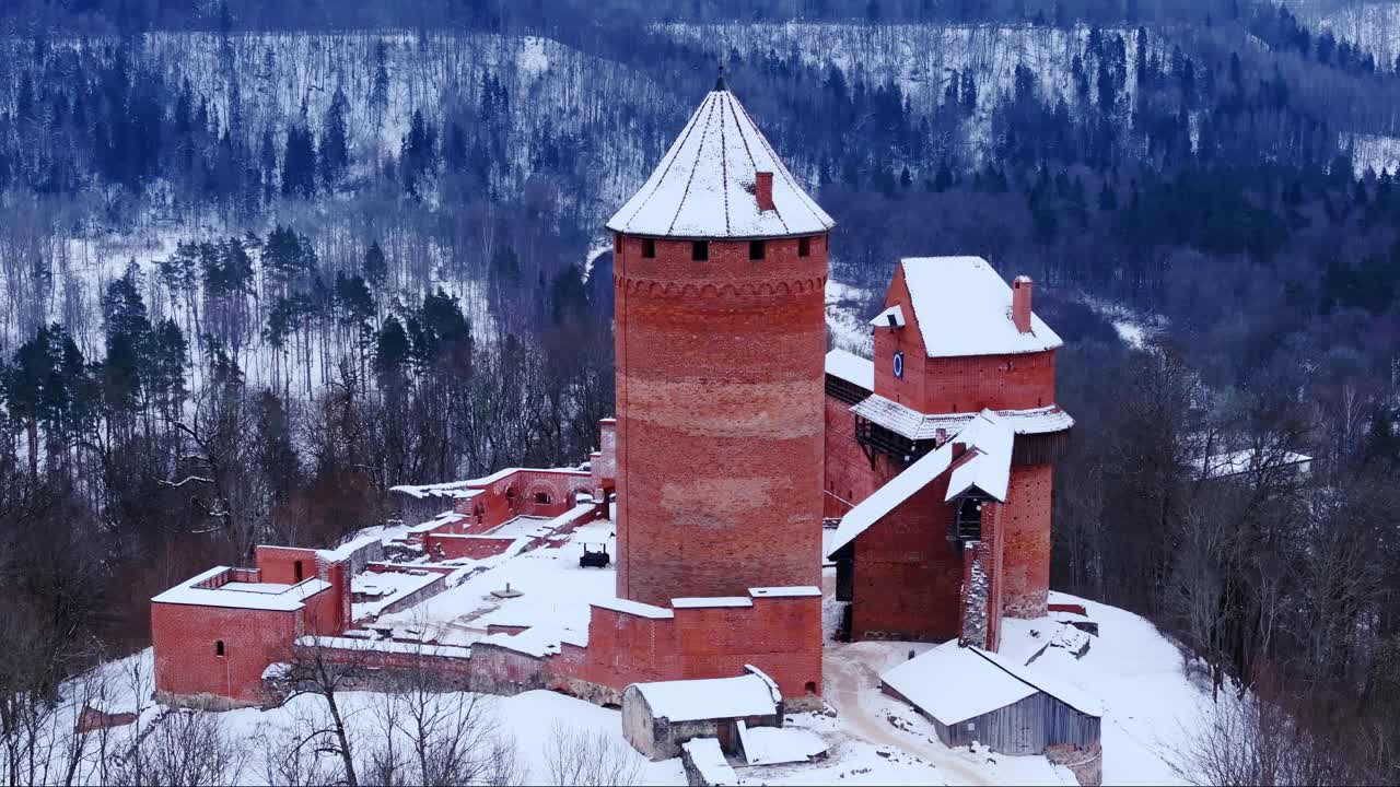 Ikonická věž z červených cihel hradu Turaida stojí na pozadí zasněžené krajiny v Lotyšsku.
