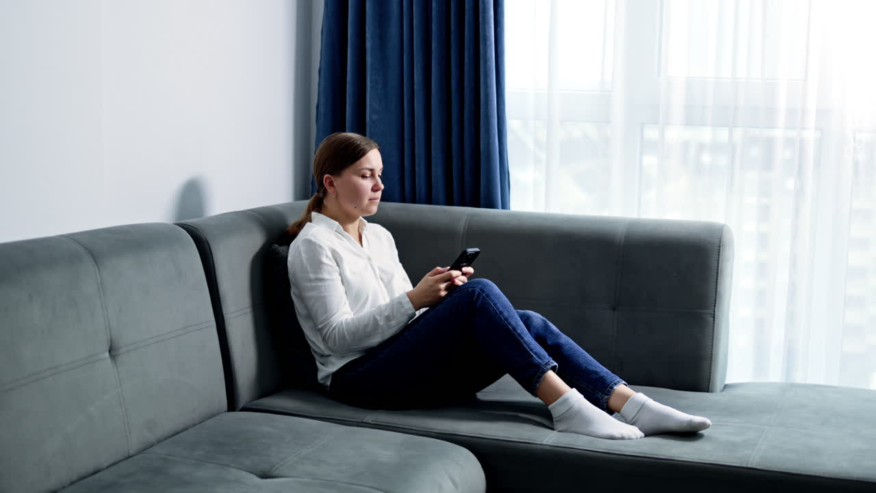 Ung kvinde i afslappet tøj arbejder online hjemme fra sofaen. En kvinde med briller og telefon i hænderne sidder i en lys stue.