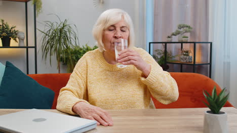 thirsty senior old woman sit at home holding glass of natural aqua make sips drinking filtered water