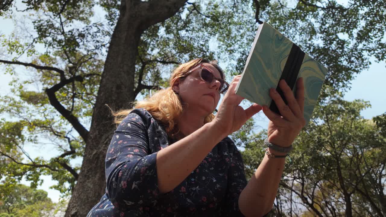 Tilt up adult caucasian woman reading book under tree. nature park. ASMR relax concept