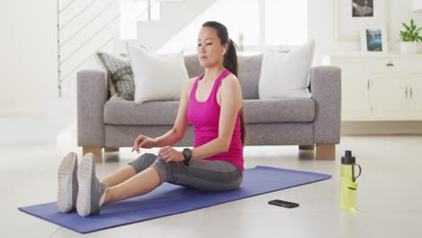 Mujer-Asiática-En-La-Alfombra-Usando-Auriculares,-Haciendo-Ejercicio-Con-Un-Teléfono-Inteligente-En-Casa