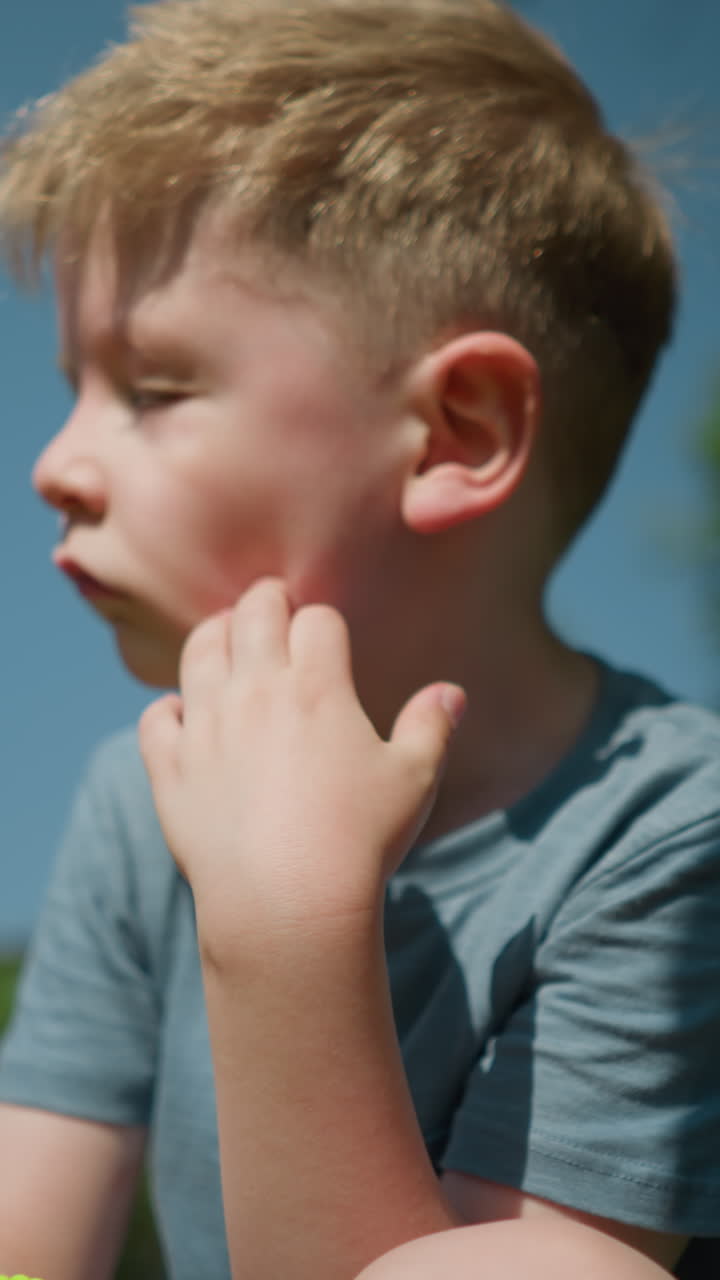 un niño pequeño con una camisa azul se frota la cara con la mano izquierda, sentado en un campo cubierto de hierba con una vista parcial de alguien cerca de él