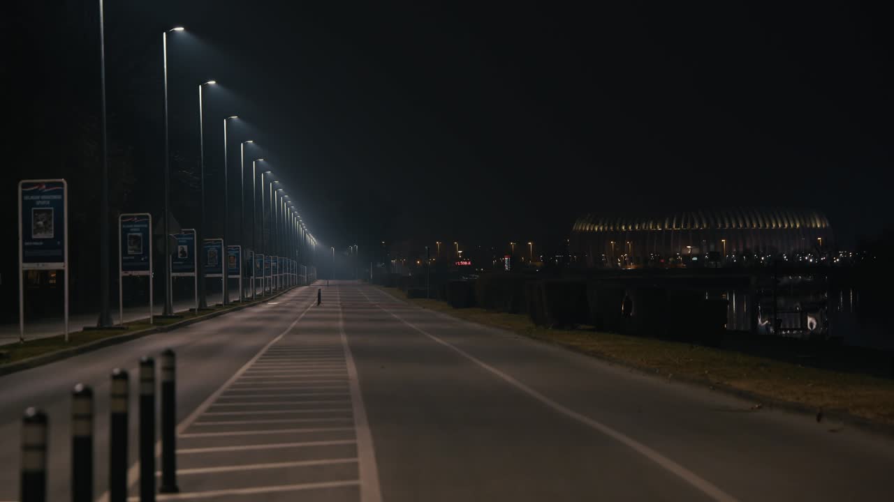 Empty road under streetlights leads towards a glowing stadium at night in Zagreb