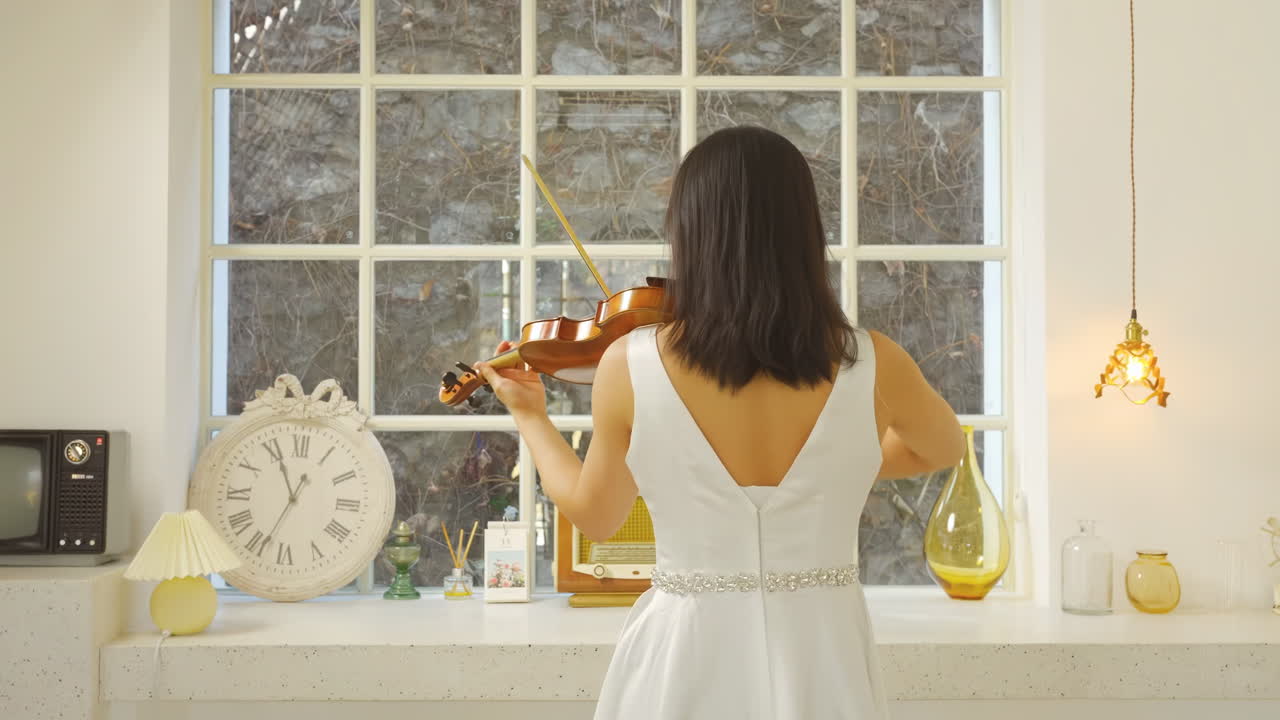 Vue de dos : femme talentueuse jouant du violon à l'ancienne, violoniste répétant sa partie avant le spectacle. Étudiante avec un violon dans une pièce lumineuse près d'une fenêtre, performance sereine, musique classique.