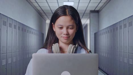 close up of asian teen girl student with a backpack smiling and typing on a laptop while standing in corridor