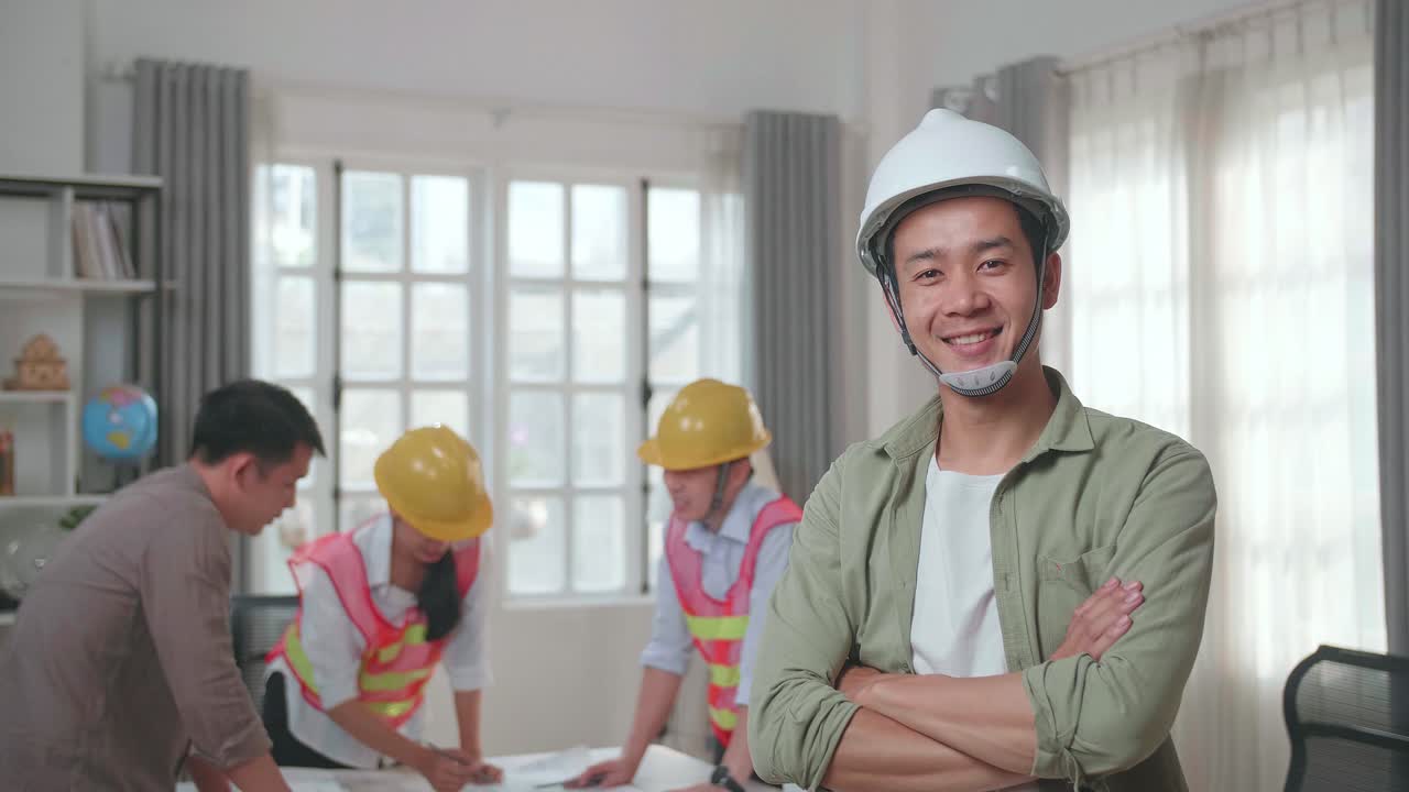 Asian Man Engineer With Helmet Crossing Arms And Smiling To Camera While His Colleagues Are Discussing About Work With A Man At The Office