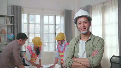 asian man engineer with helmet crossing arms and smiling to camera while his colleagues are discussing about work with a man at the office