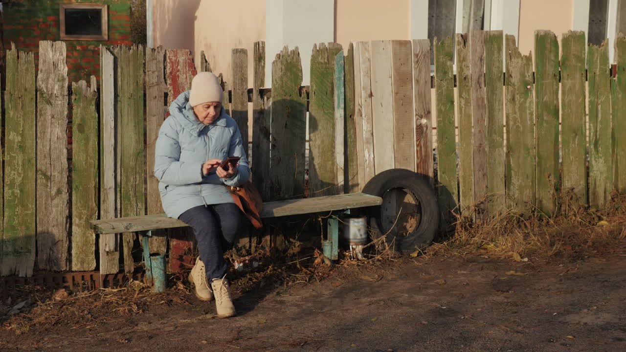старшая женщина с мобильным телефоном на скамейке у деревянного забора