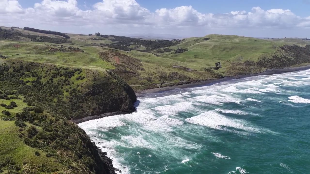 Ripresa con drone in avvicinamento su dolci colline verdi verso una spiaggia di sabbia nera e onde turchesi dell'oceano, Nuova Zelanda, vista paesaggistica epica.