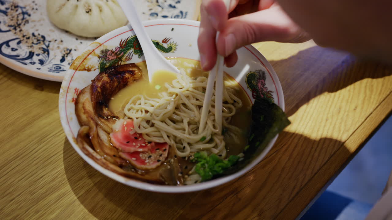 Mano che solleva noodles da una ciotola di ramen con le bacchette, mentre è seduto a un tavolo.