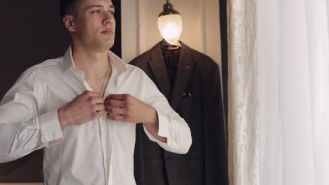 stylish man in jacket fastens buttons, buttoning white shirt preparing to go out near window