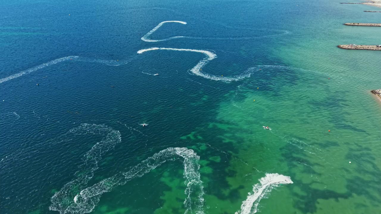 Jet ski racing takes place on a clear day in open waters with waves and trails left behind by the speeding jet skis