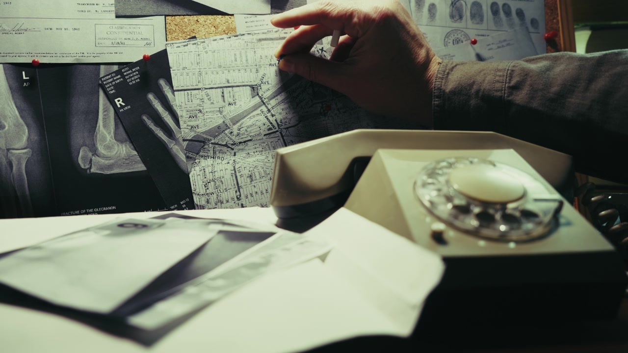 Una mano appunta nuove prove su una bacheca investigativa ingombra, circondata da documenti e un telefono retrò, suggerendo lavoro investigativo, analisi criminale e la ricerca di connessioni nascoste