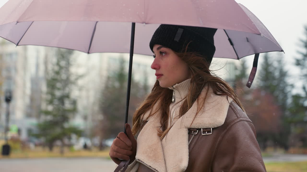 Female holding umbrella wearing black knit cap and brown shearling jacket, surrounded by pine trees and urban buildings, soft snowfall falling on cold overcast day