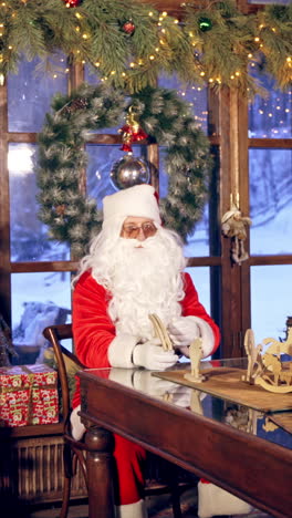 santa claus is sitting in specially decorated room at the table, holding wooden dear near the new year tree. christmas spirit concept close up video. christmas eve. new year is coming. vertical video