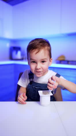 lovely toddler boy in jeans romper sits at big dinner table in the kitchen. cute kid is eating yogurt. vertical video.