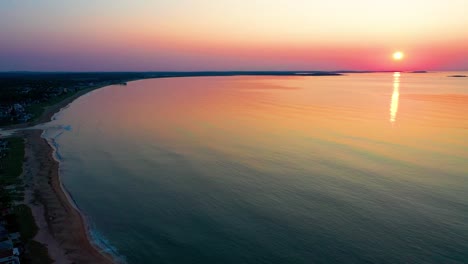 Vista-Panorámica-Izquierda-Del-Hermoso-Amanecer-En-La-Playa-Con-Un-Sol-Brillante-Que-Proyecta-Un-Colorido-Reflejo-Rojo,-Naranja,-Púrpura-Y-Amarillo-Sobre-Las-Casas-De-Vacaciones-Y-Las-Ondulantes-Olas-Del-Océano-Y-El-Hermoso-Cielo