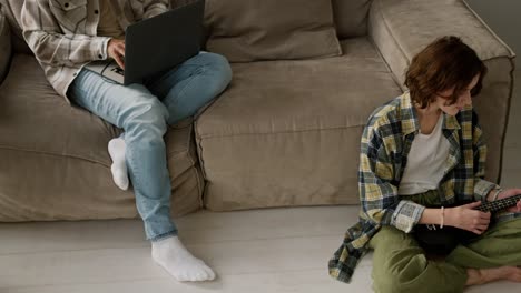 a girl with brown hair with a bob hairstyle in a checkered shirt plays the ukulele while her boyfriend a young brunette with black skin color in black headphones works on his gray laptop sitting on the sofa in a modern studio apartment