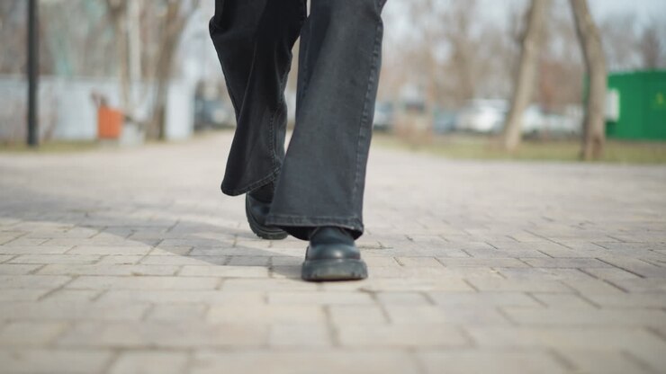 Nahaufnahme aus der Untersicht einer Frau, die an einem sonnigen Tag im Stadtpark auf einem gepflasterten Weg vorwärtsgeht, schwarze Schuhe und weite Jeans tragend, was Bewegung, Mode und einen aktiven Outdoor-Lebensstil hervorhebt.