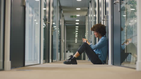 young businesswoman sitting on floor in corridor of modern office with phone celebrating good news