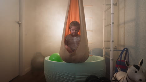 Un-Niño-Frunce-El-Ceño-Sentado-En-Una-Hamaca-Naranja-Encima-De-Una-Piscina-De-Bolas