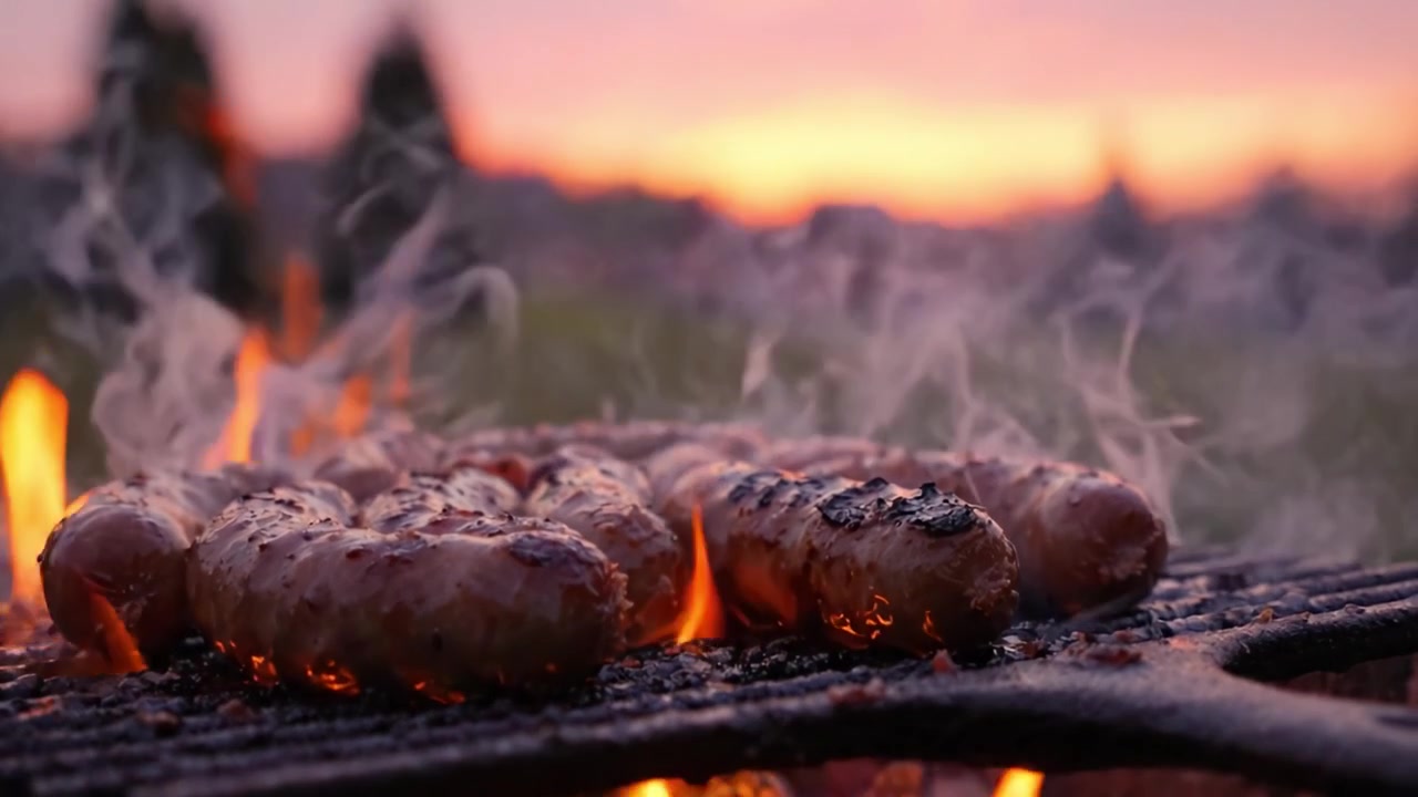 Affascinante scena di barbecue che mostra salsicce sfrigolanti su una griglia, circondate da fiamme vivaci e uno sfondo di tramonto colorato che esalta l'atmosfera della cottura.