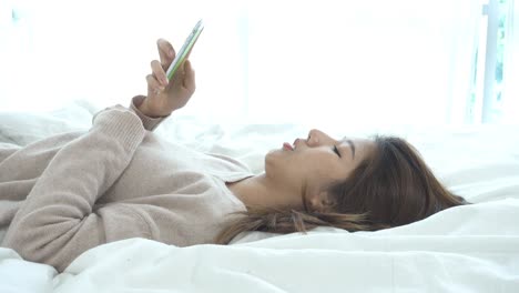 행복한 아시아 여성은 아침에 침대에서 스마트 폰을 사용합니다. 아시아 여성은 침대에서 스마트폰으로 소셜 앱을 확인합니다. 웃는 여성은 집에서 드폰으로 인터넷을 서핑합니다. 모바일 중독자 개념.