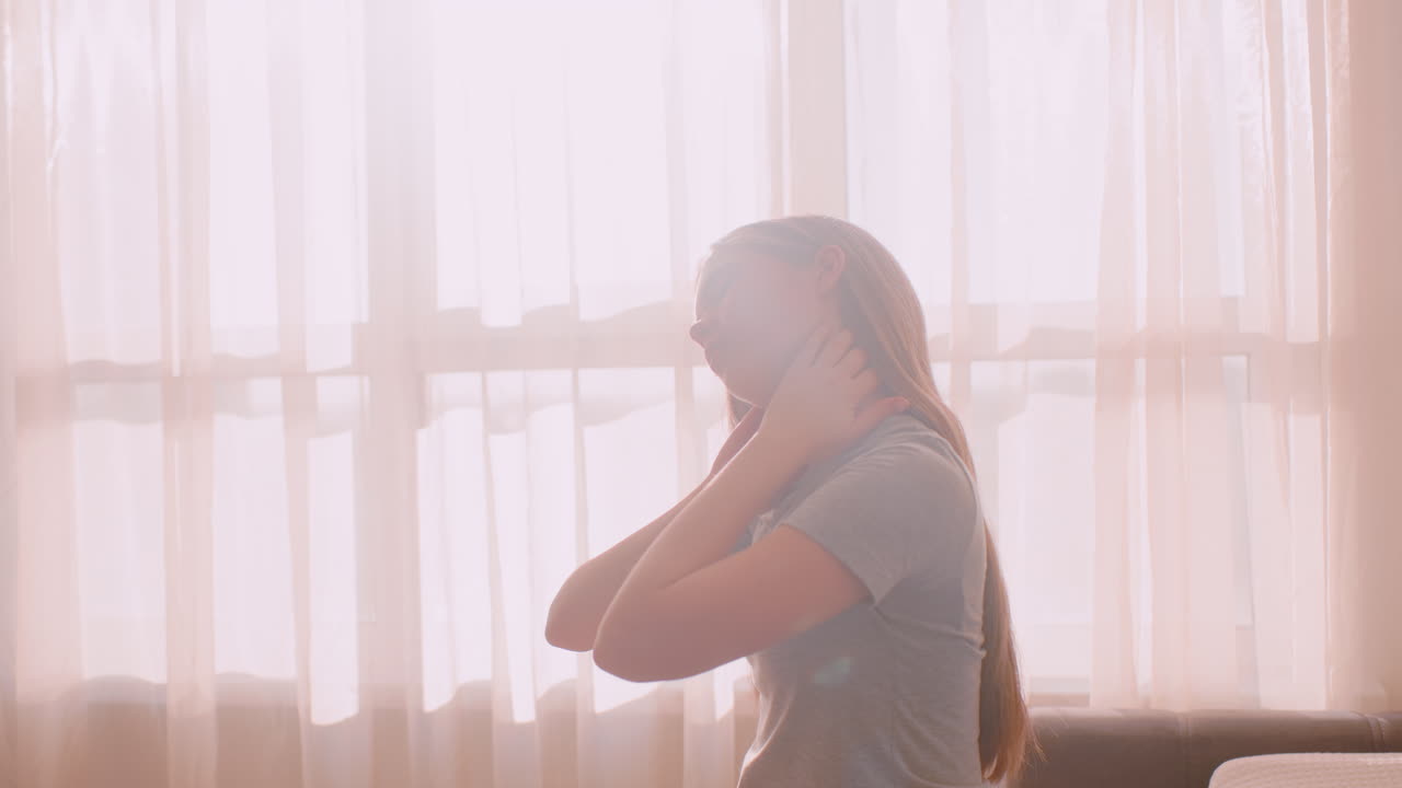 Side view of young woman with long hair stretching neck and massaging nape with both hands while s in front of sunlit curtain, expressing tension relief