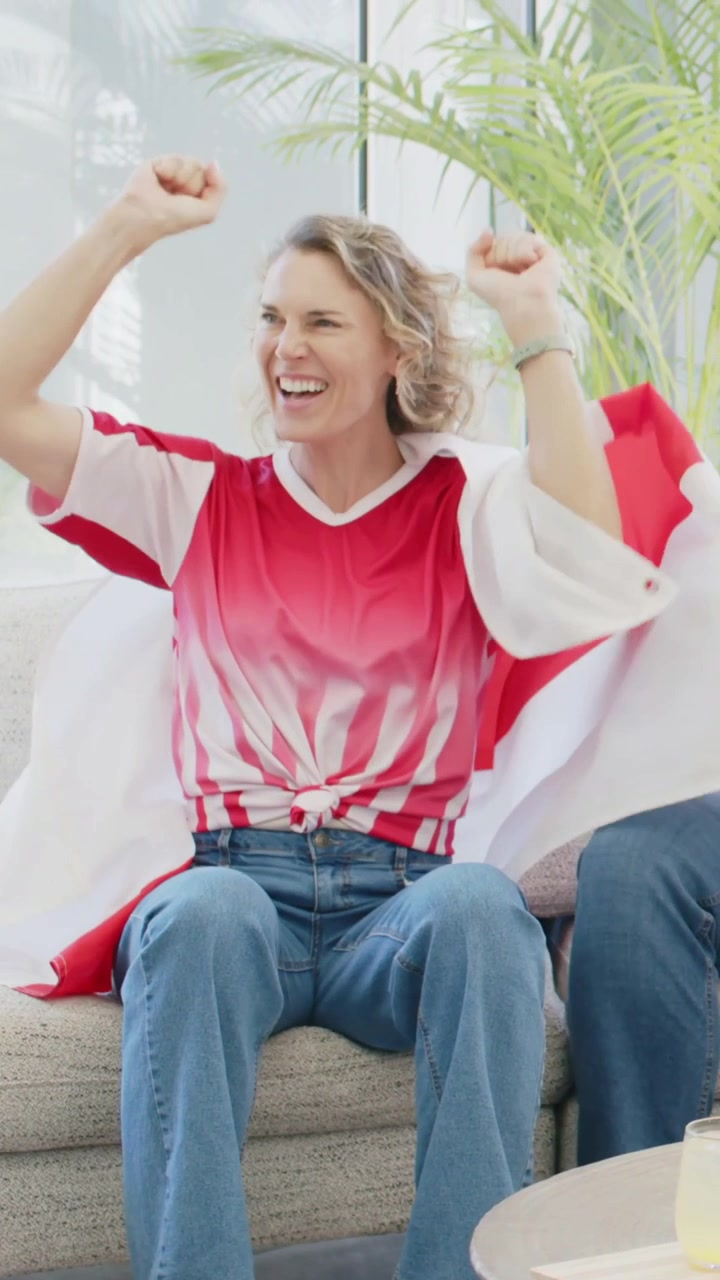 Vertical video: Clapping, woman in red white shirt and flag cheering on sofa after offscreen event