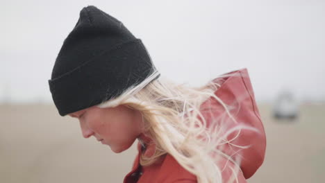 young girl wearing black beanie and red hooded jacket looking down while the wind blows her blonde hair in muiden, netherlands