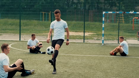 young street football player training freestyle tricks while two team players sitting on the pitch doing stretch exercises and another one using mobile phone 1