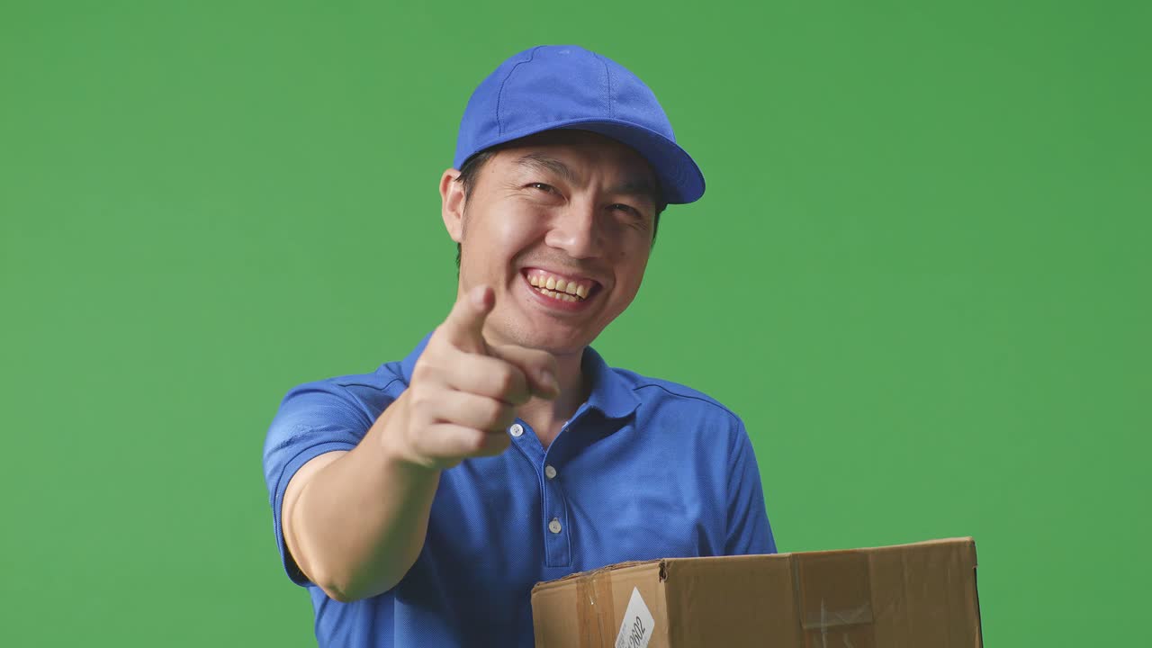 푸른 유니폼을 입은 아시아 남성 택배기사의 클로즈업은 미소 짓고 스튜디오에서 녹색 스크린 배경에 상자를 배달하는 동안 정직한 몸짓을합니다.