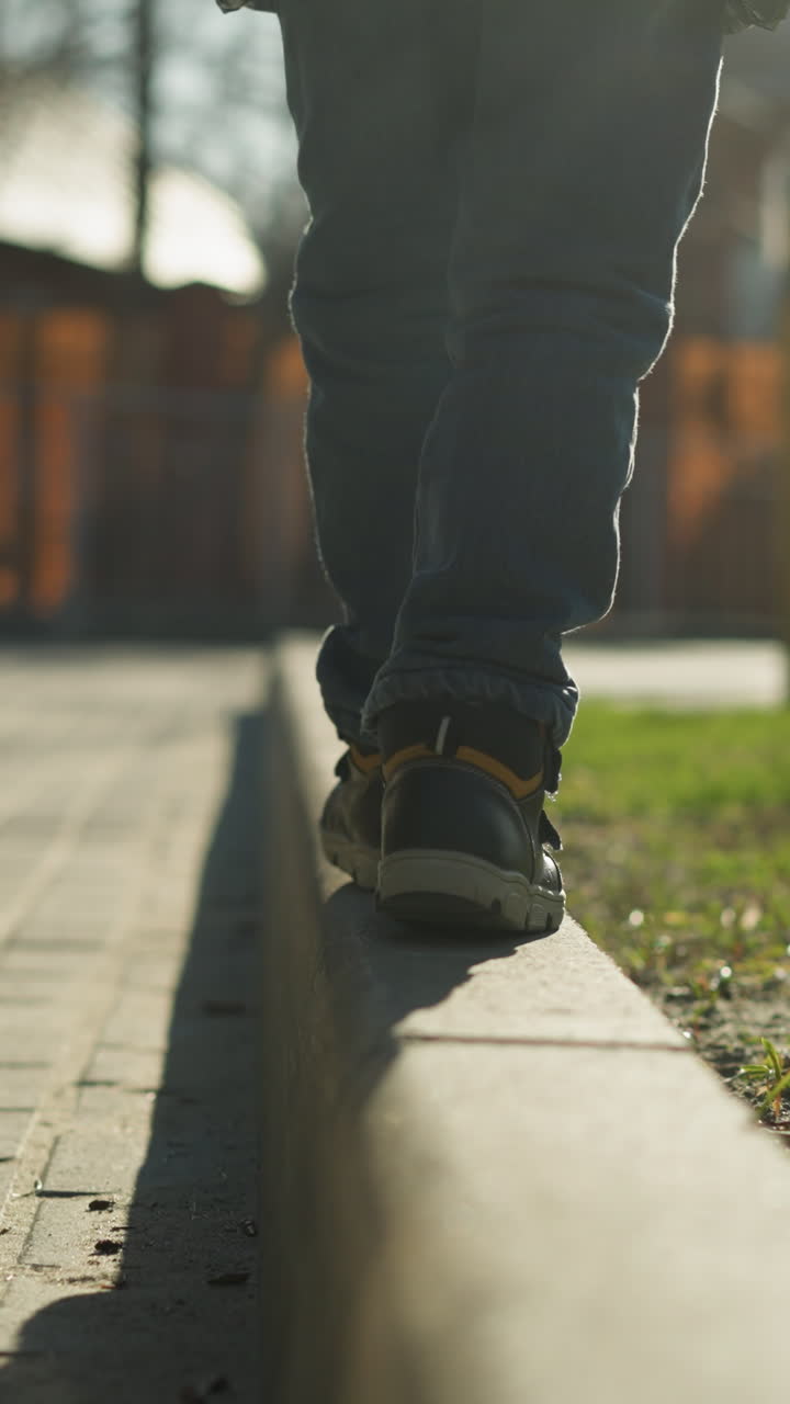 un primer plano de un niño equilibrado cuidadosamente mientras camina a lo largo de un borde del pavimento al lado de un coche, vestido con una chaqueta negra brillante, con una vista borrosa de árboles y edificios en el fondo
