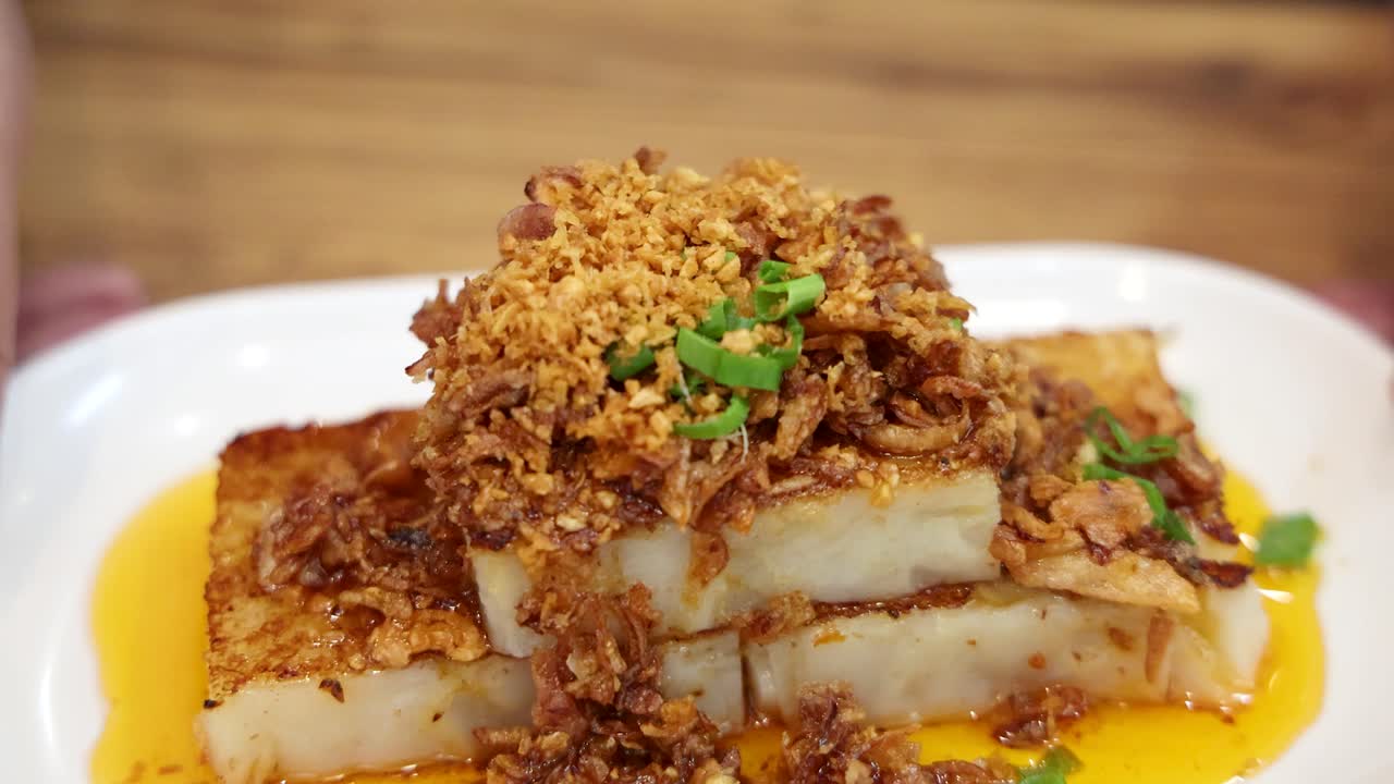 Hands present a plate of fried turnip cake topped with crispy shallots and green onions under bright, even lighting on a wooden table, camera moves in smoothly