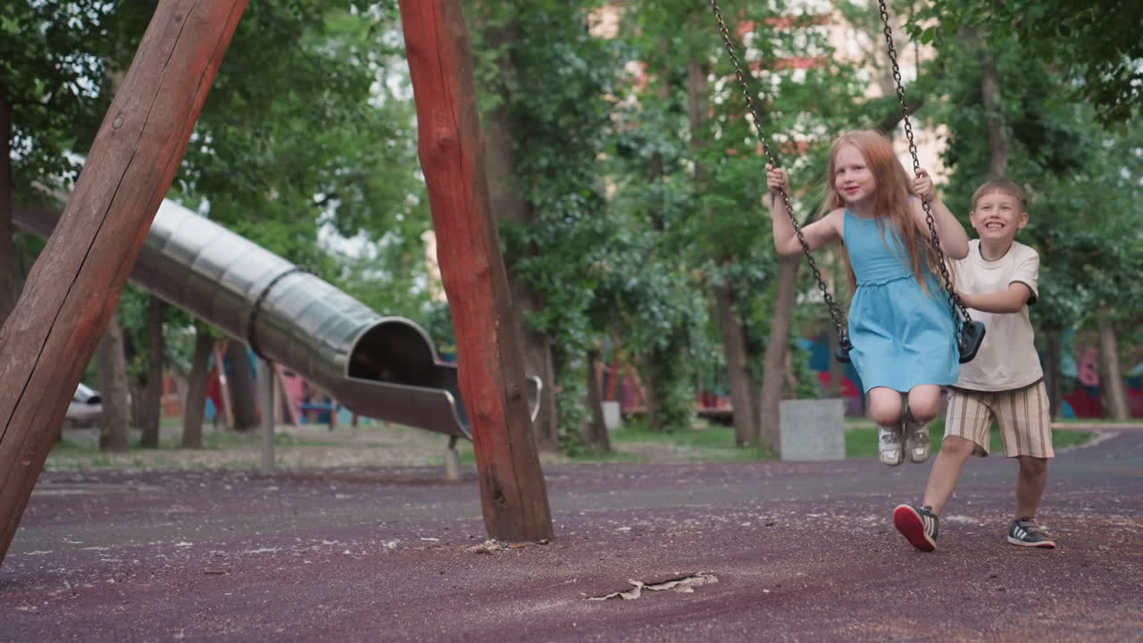 playful girl laughs out loud on wooden swing with chain handle as her playmate pushes from behind in lush green park, capturing candid joy motion blur and sibling fun in warm evening light