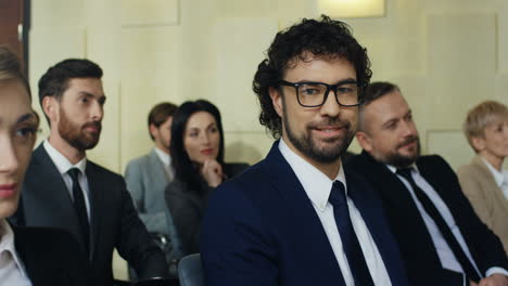 vista de cerca de un hombre de negocios caucásico con anteojos sentado entre la gente en una sala de conferencias, luego gira su rostro hacia la cámara y sonríe