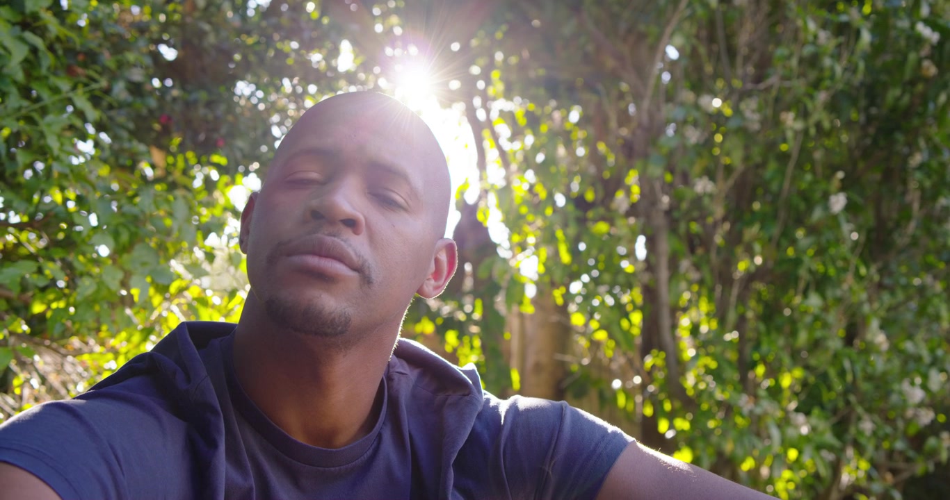 African American adult male wearing hoodie by fence noticing sun flare shifting gaze facing camera