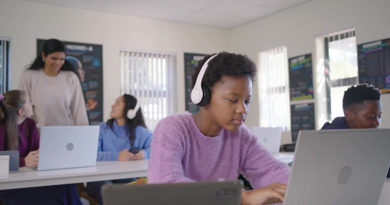 Insegnante che passa, compagni di classe che reagiscono, adolescente afroamericano che digita su dispositivi alla scrivania mentre lavora.