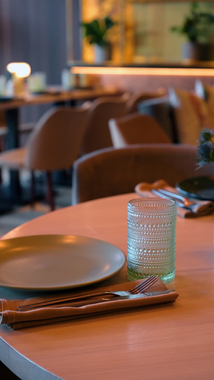 Minimalist table setting in restaurant with green glass. Close up of a wooden table set with a textured green glass, beige plate, and cutlery in a napkin