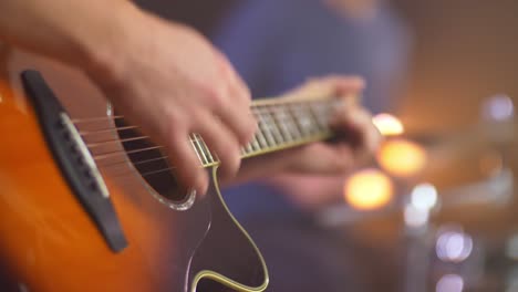 Primer-Plano-De-Un-Músico-Profesional-Tocando-La-Guitarra-Occidental-Con-Un-Baterista-Borroso-En-El-Fondo-Durante-Una-Sesión-En-Vivo