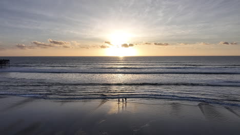 Disparo-De-Un-Dron-Orbitando-Alrededor-De-Dos-Personas-Caminando-En-La-Costa-Del-Océano-Al-Atardecer