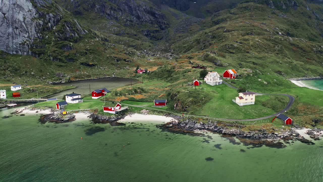 vista aérea, órbita inclinada hacia arriba, unas pocas casas de rorbu rojas en un pequeño pueblo pesquero costero al pie de majestuosas montañas en las islas loftoten, noruega
