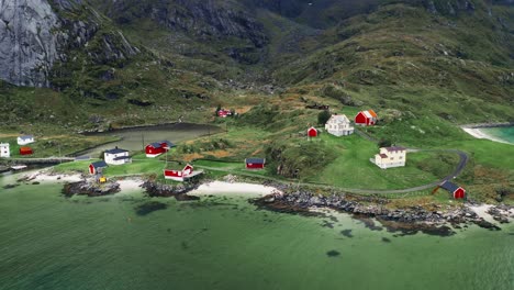 aerial view, orbit tilt-up, a few red rorbu houses in a tiny coastline fishing village at the foot of majestic mountains in the lofoten islands, norway