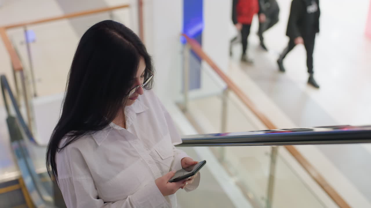 Confident woman wearing glasses and white shirt turns while holding phone on escalator in brightly lit mall interior with people in motion and retail background in view